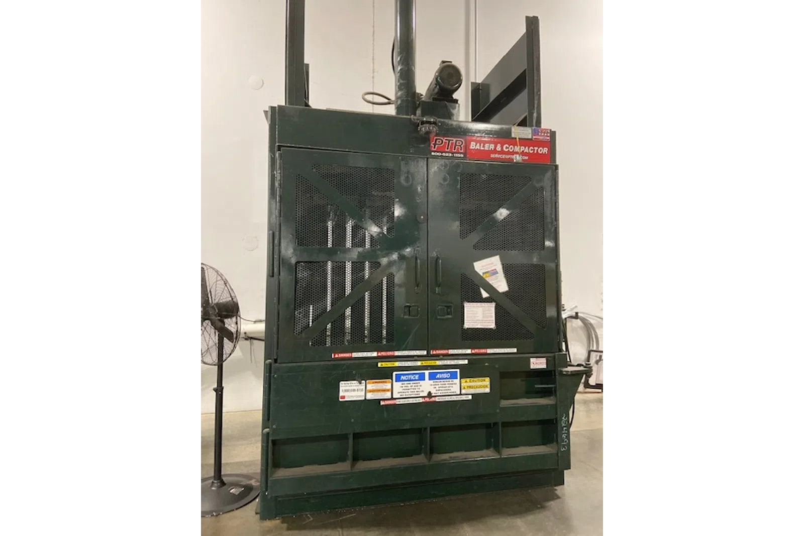 Industrial PTR baler and compactor machine in warehouse setting with caution labels and metal mesh doors.