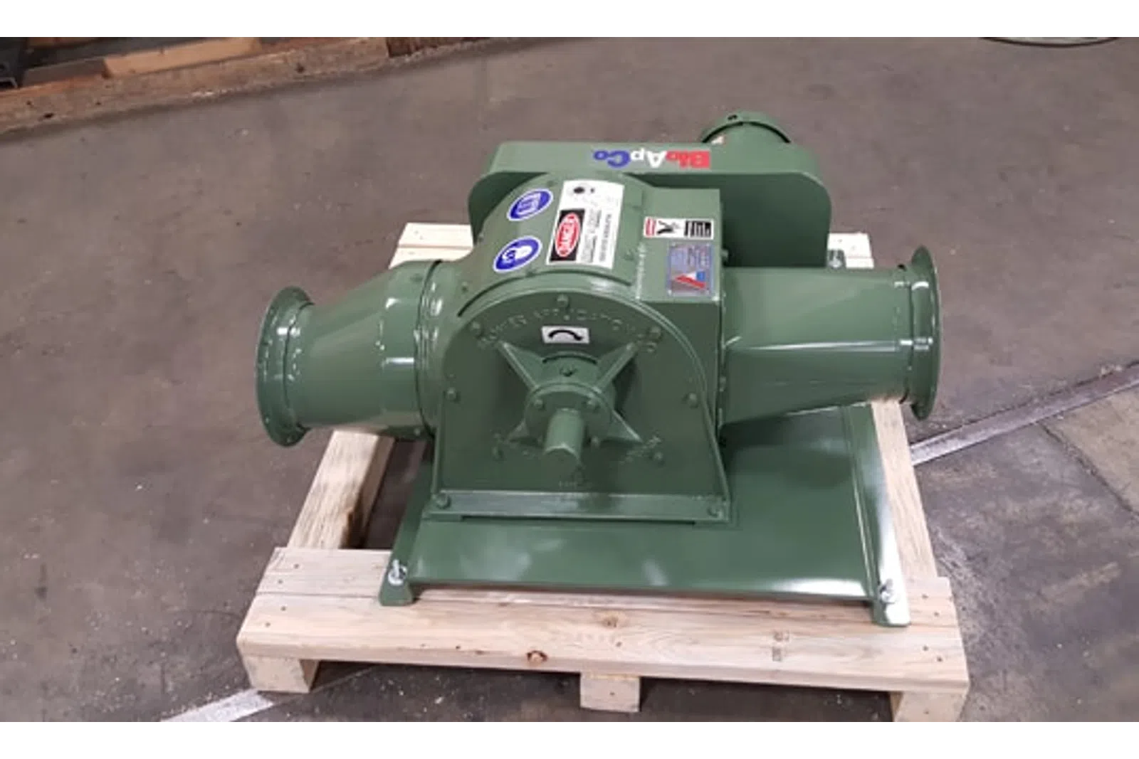 Green industrial blower mounted on a wooden pallet inside a workshop with concrete floor.