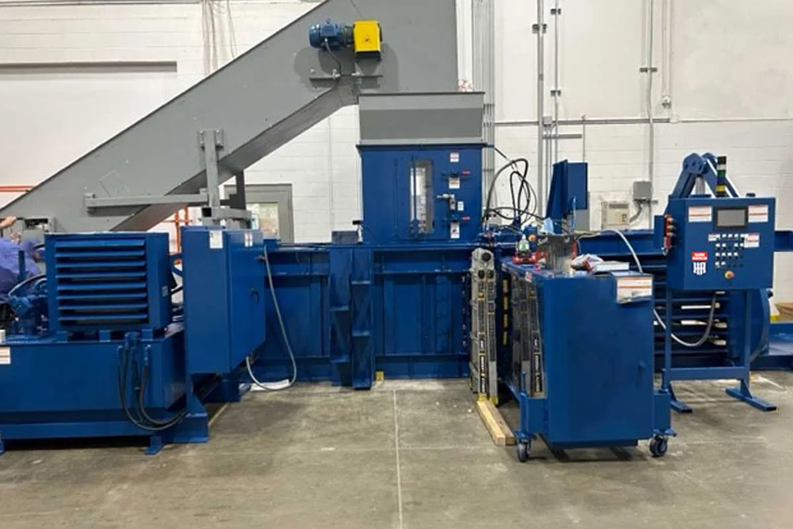 Large blue industrial baler machine with conveyor belt in a factory setting on concrete floor.