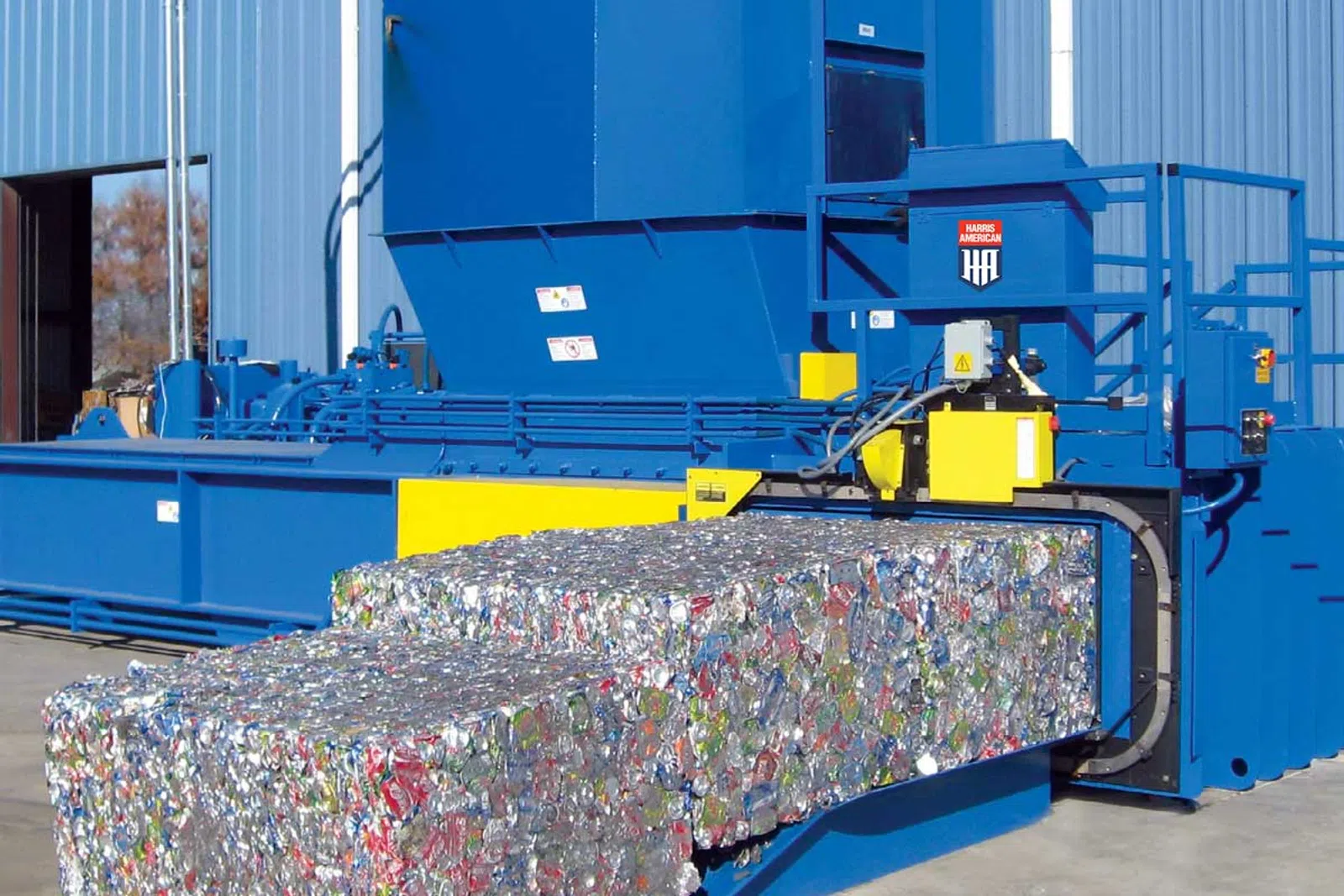 Industrial blue recycling baler compresses crushed aluminum cans into large compact bales outdoors.