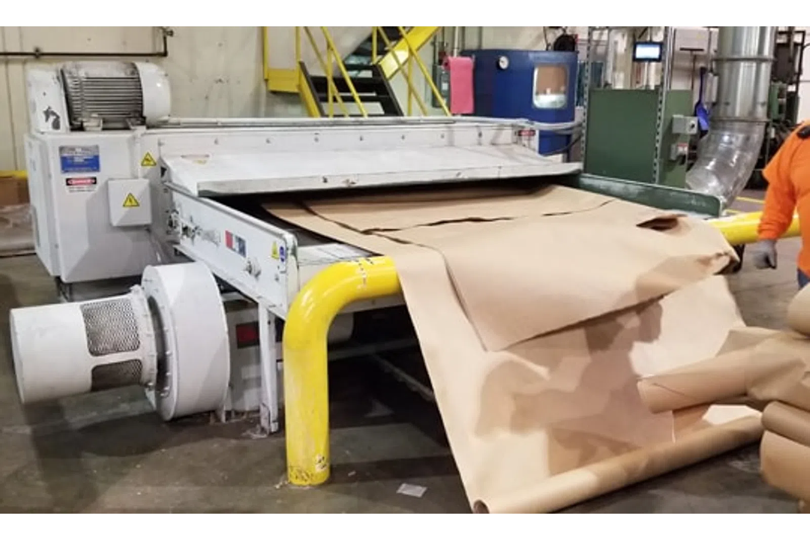 Industrial machine processing large rolls of brown kraft paper in a factory setting with yellow safety railings.