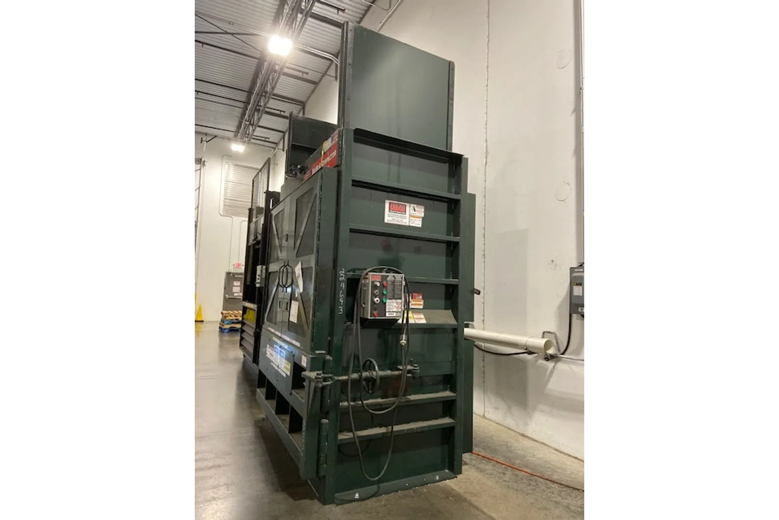 Large green industrial machine with control panel and cables in a warehouse with concrete floor and high ceiling.
