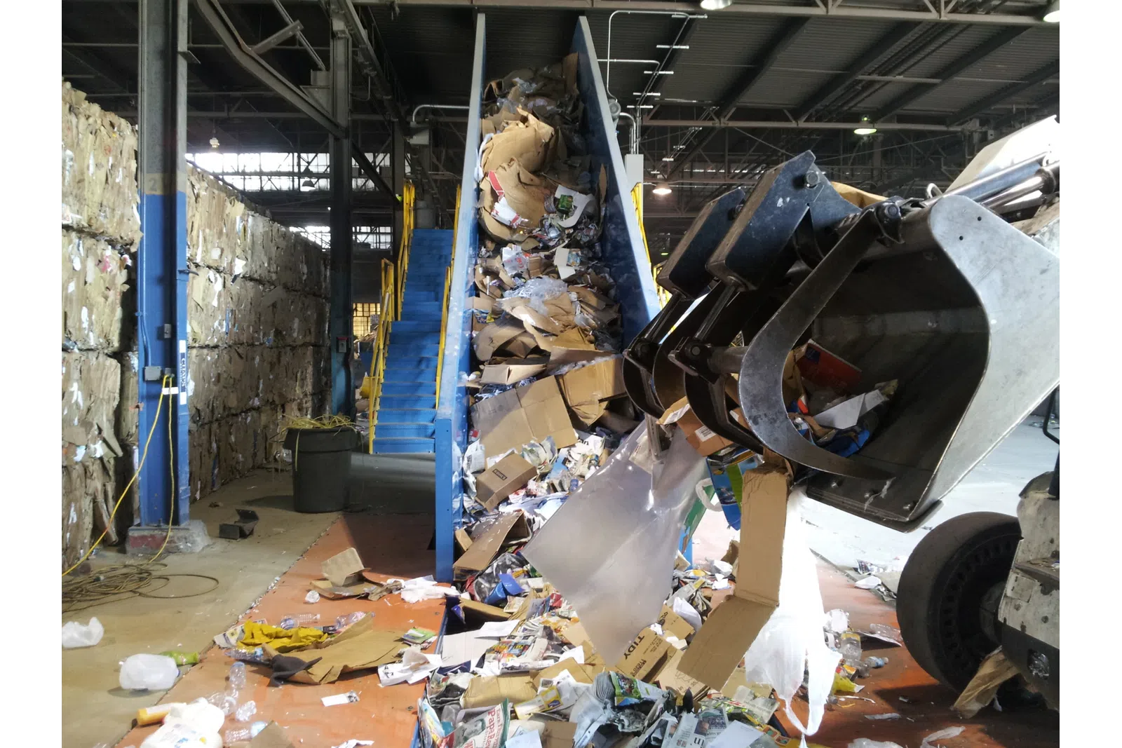 Recycling facility conveyor belt loaded with cardboard and paper waste being handled by industrial machinery.