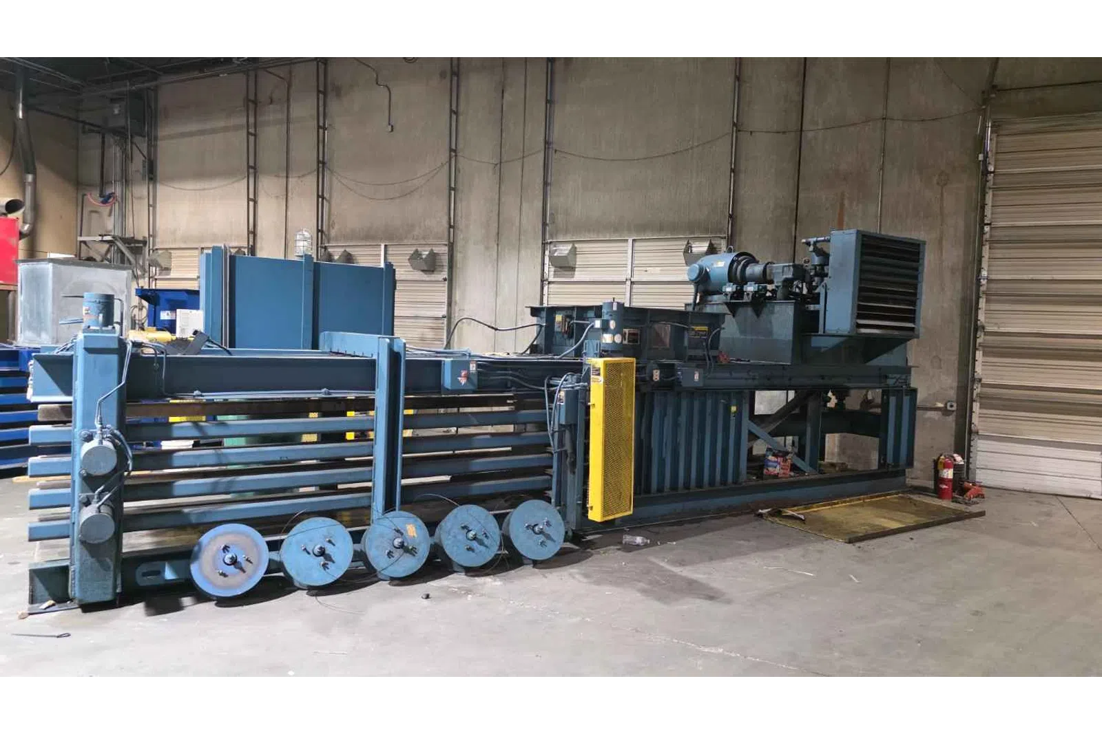 Large industrial baler machine with multiple rollers inside a warehouse with concrete walls and garage doors.