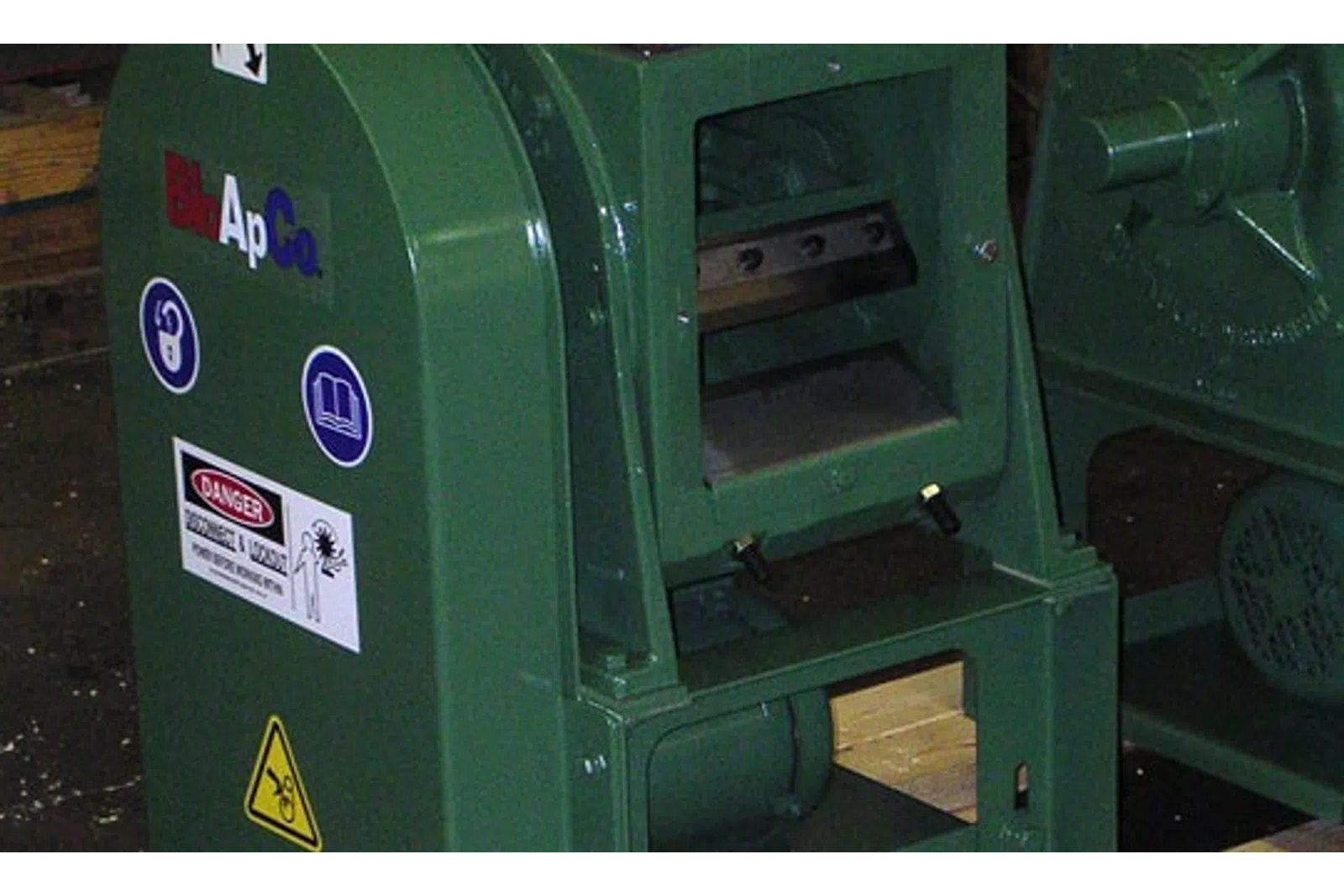 Green industrial machine with warning labels and safety signs in a workshop setting.
