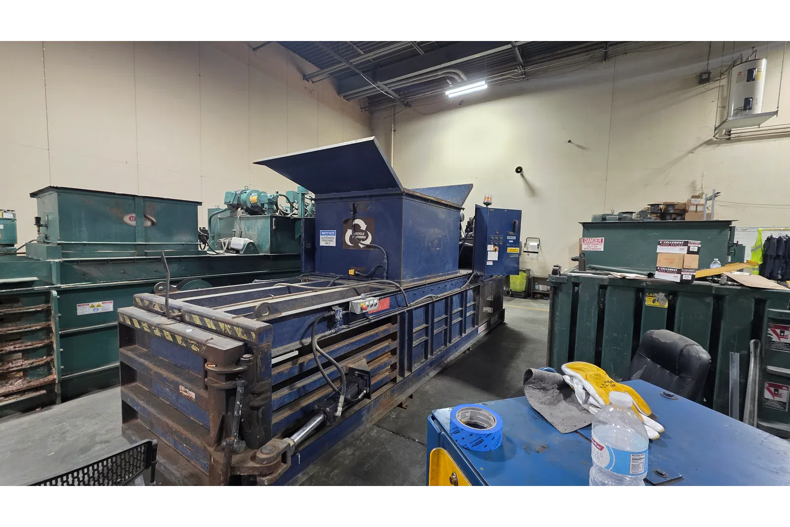 Industrial warehouse with large blue metal baler machine and nearby recycling bins in a sorting facility.