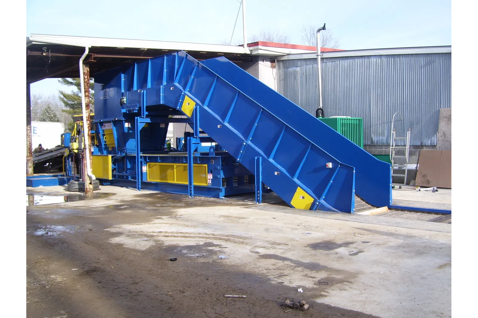 Large blue industrial machine with conveyor belt outdoors next to warehouse building on concrete ground.