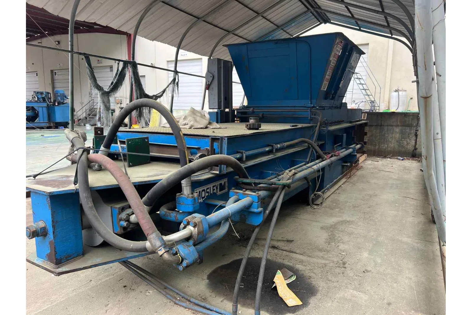 Large industrial blue metal machine with hoses inside a covered workspace on concrete floor.