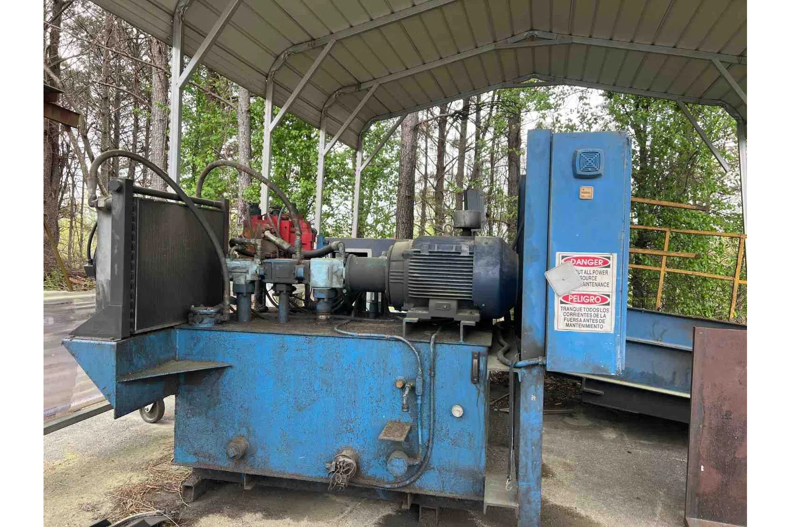 Industrial blue hydraulic machine with electric motor under a metal canopy outdoors surrounded by trees.