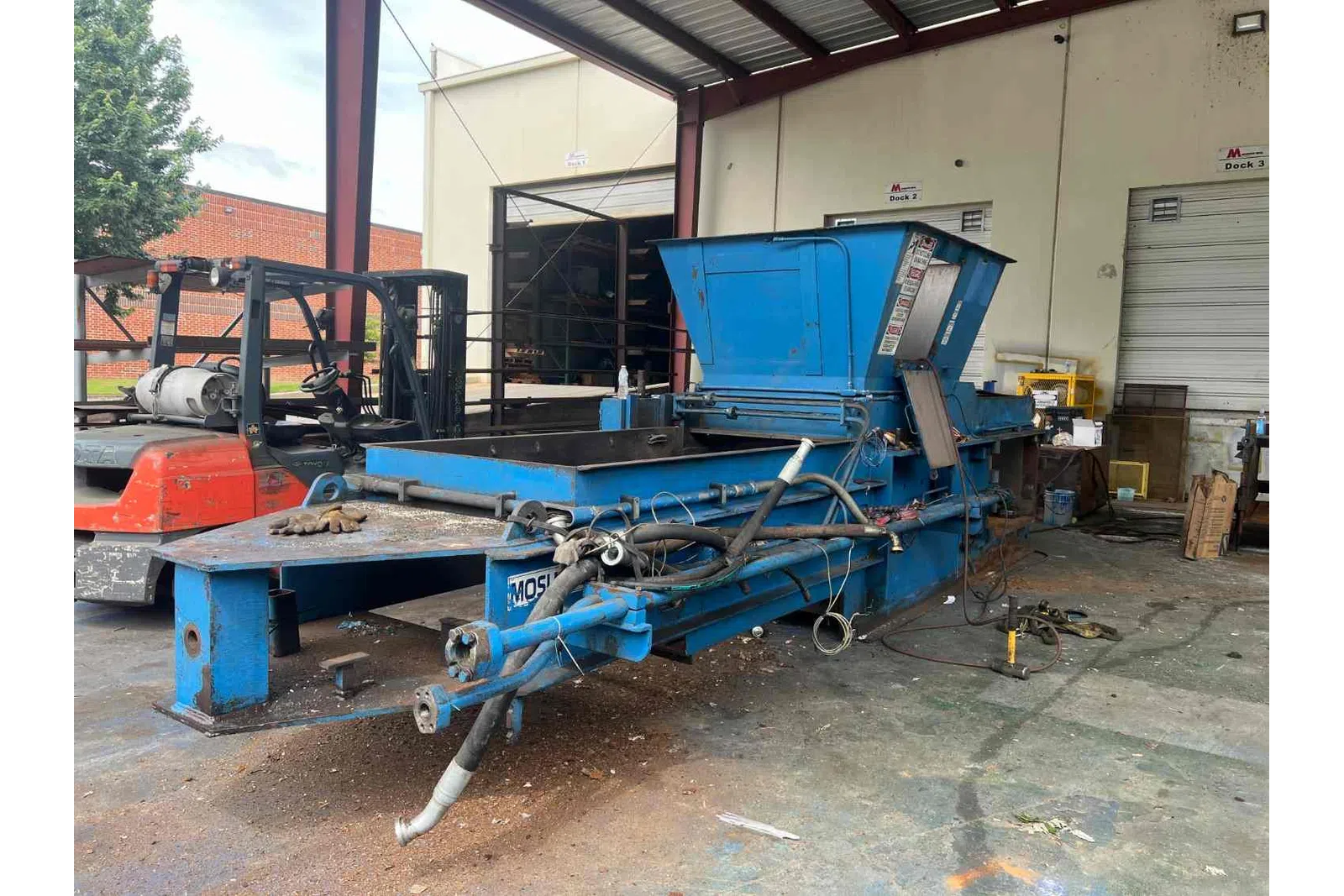 Industrial blue metal equipment with hoses and pipes inside warehouse near forklift and loading docks.