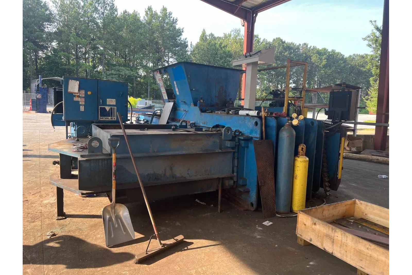 Industrial metal baler machine with gas cylinders under a metal roof in an outdoor workshop area.