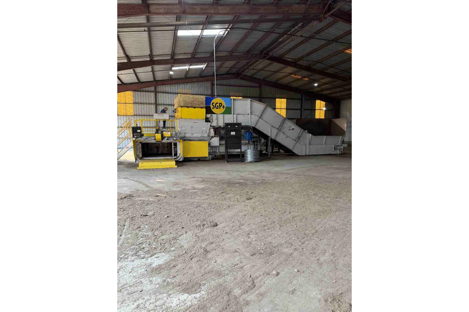 Industrial processing machine with yellow and grey parts inside a large warehouse with metal roof beams.