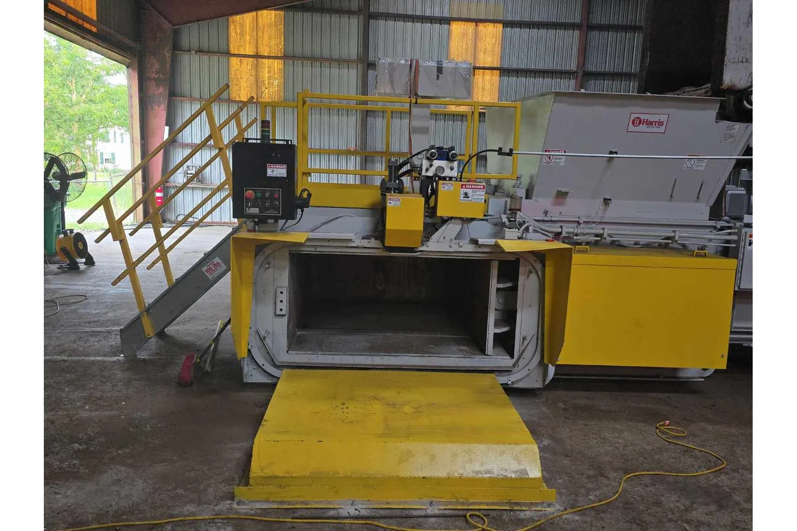 Industrial yellow waste compactor machine inside a warehouse with safety railings and control panel.