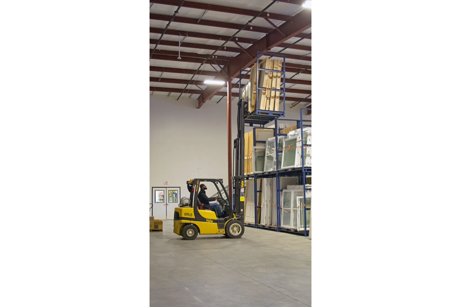 Man operating yellow Yale forklift lifting wooden panels in a spacious warehouse with organized shelves.