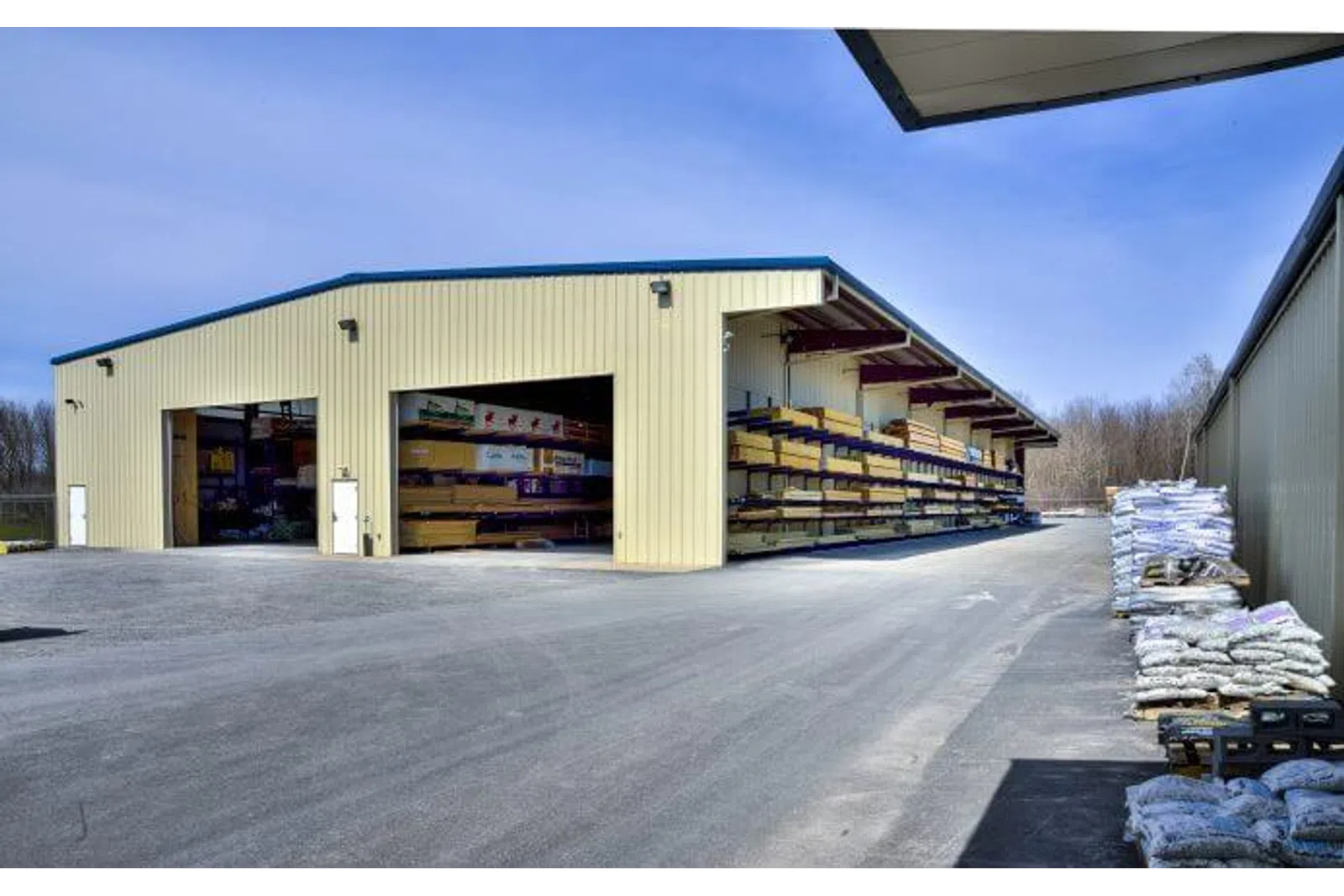 Large metal warehouse with open storage racks holding lumber and piles of bags outside on paved ground under blue sky