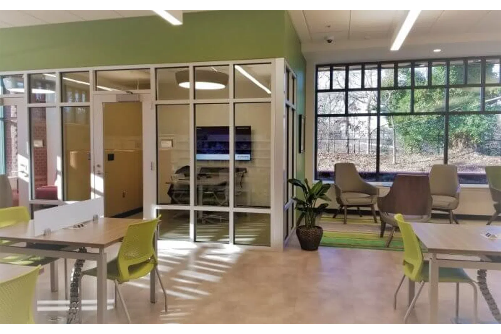 Modern office space with glass meeting room, green chairs, large window, and natural light filled room.