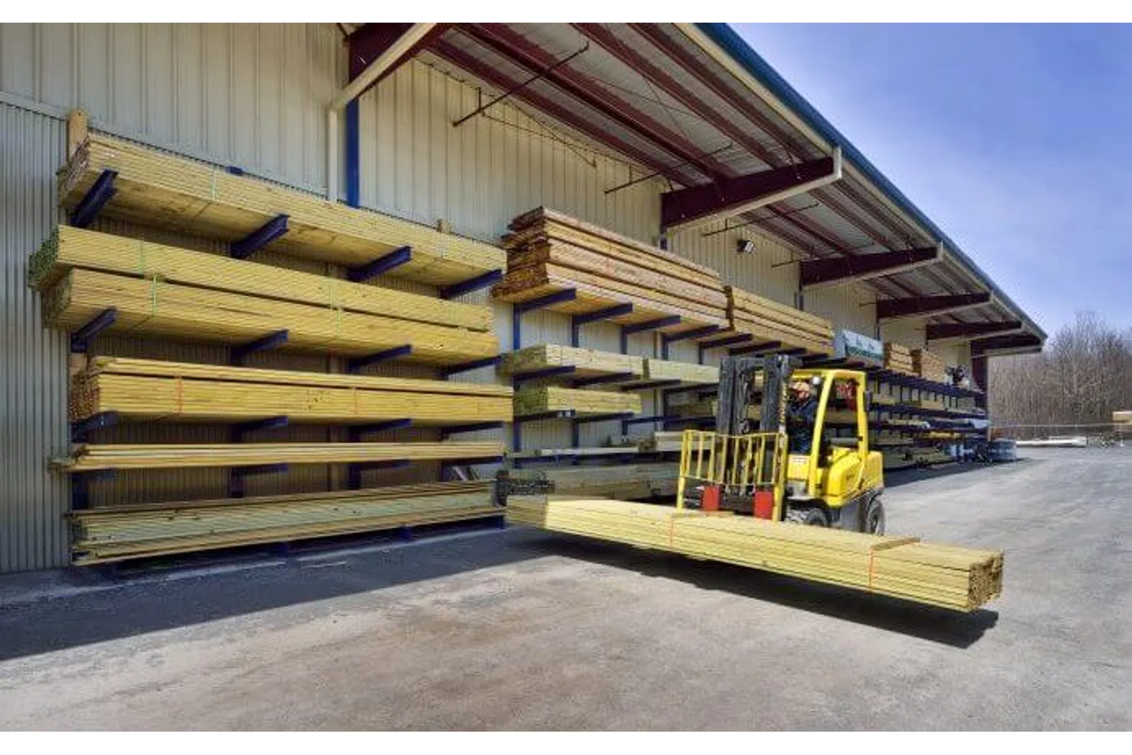 Yellow forklift carrying long wooden planks outside a lumber store with stacks of wood on metal racks under roof.