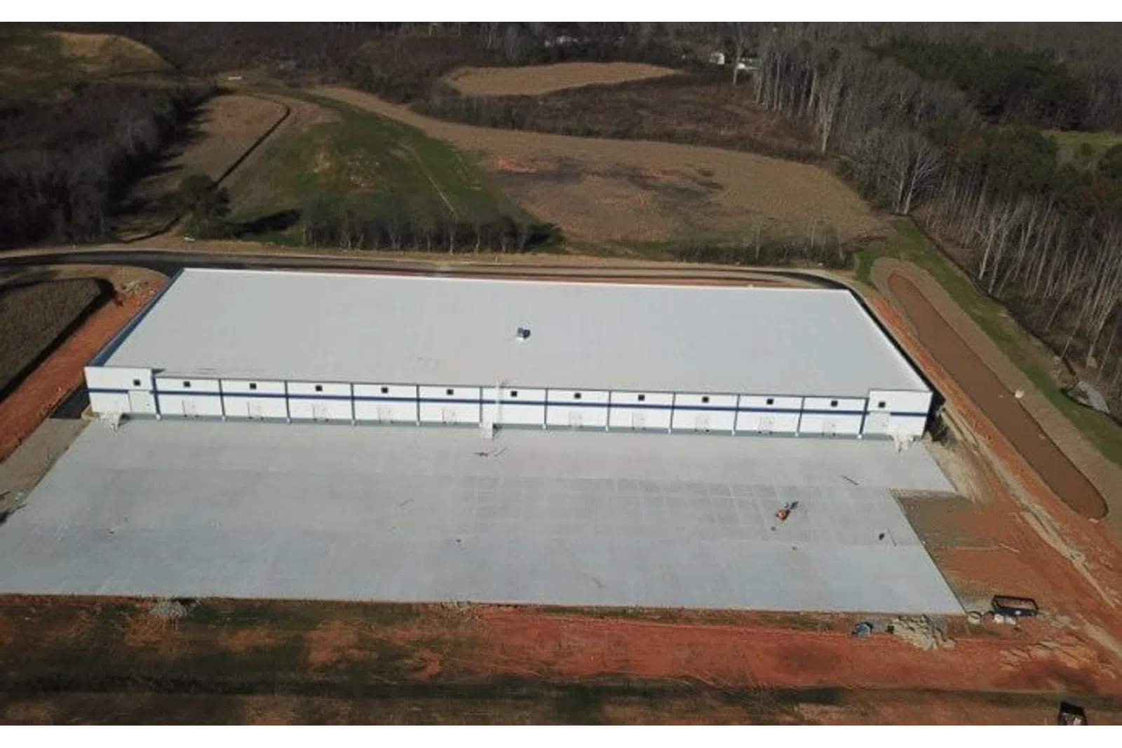 Aerial view of a large new industrial warehouse with a white roof surrounded by cleared land and forest.