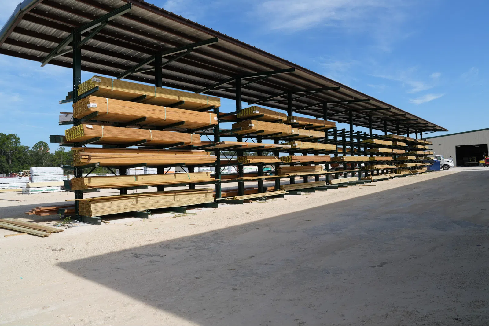 Outdoor lumber yard with stacked wooden planks under a metal canopy on a sunny day