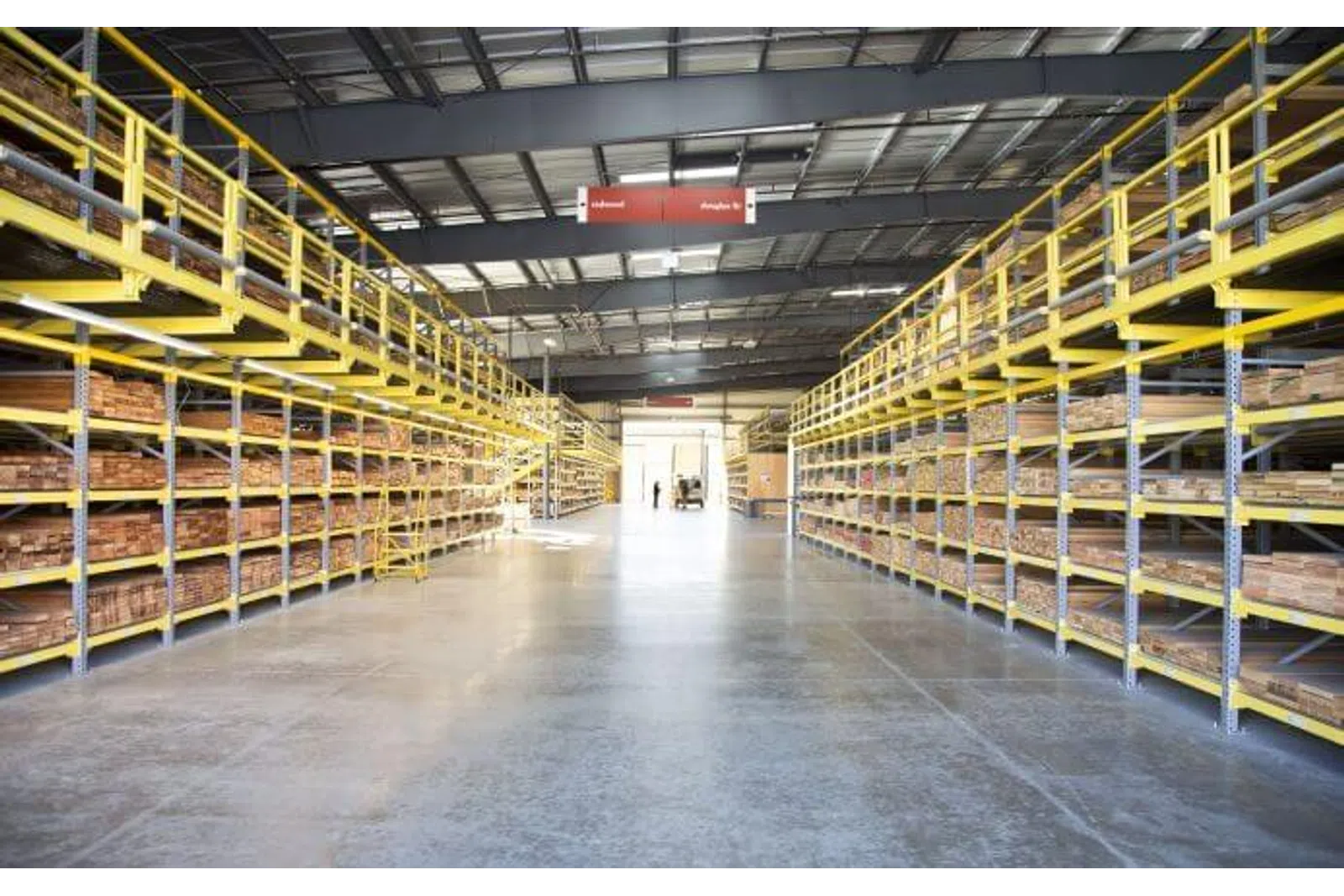 Spacious warehouse with yellow industrial shelving stocked with wooden planks under a metal roof with bright lighting.