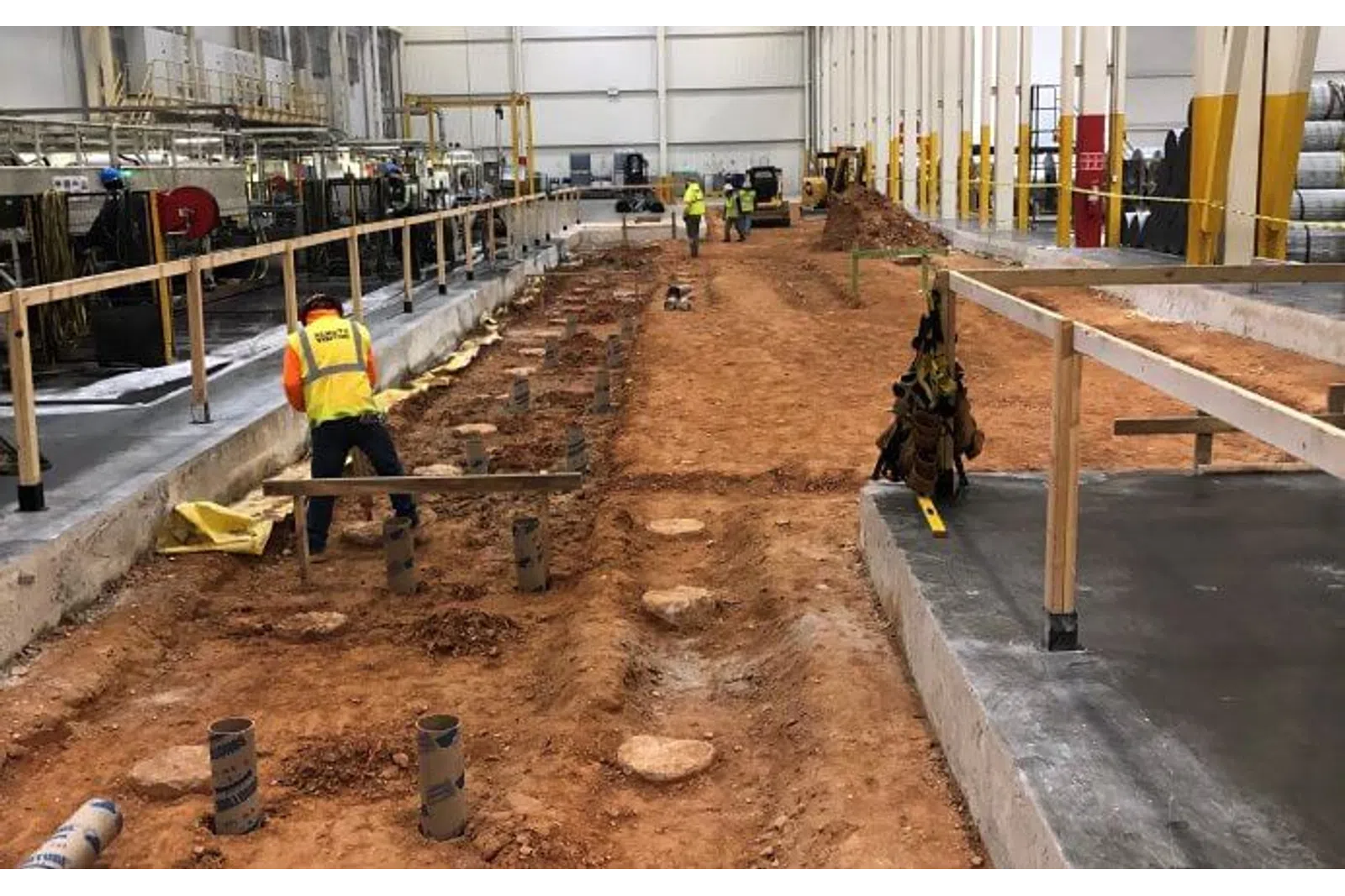 Construction workers preparing a large industrial site foundation with soil excavation and concrete flooring inside a warehouse.