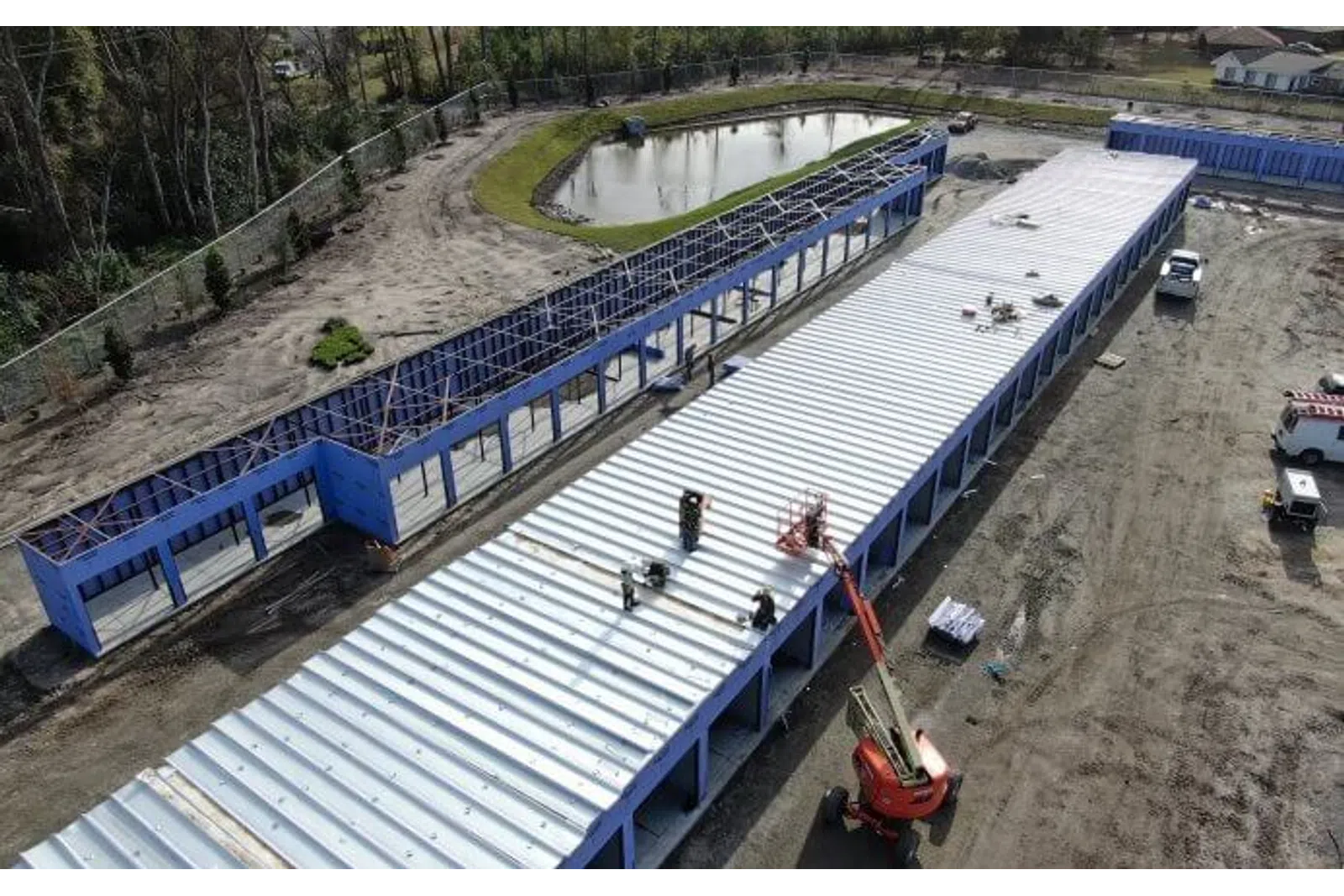 Aerial view of blue-framed industrial buildings under construction near a pond with workers on the metal roof.
