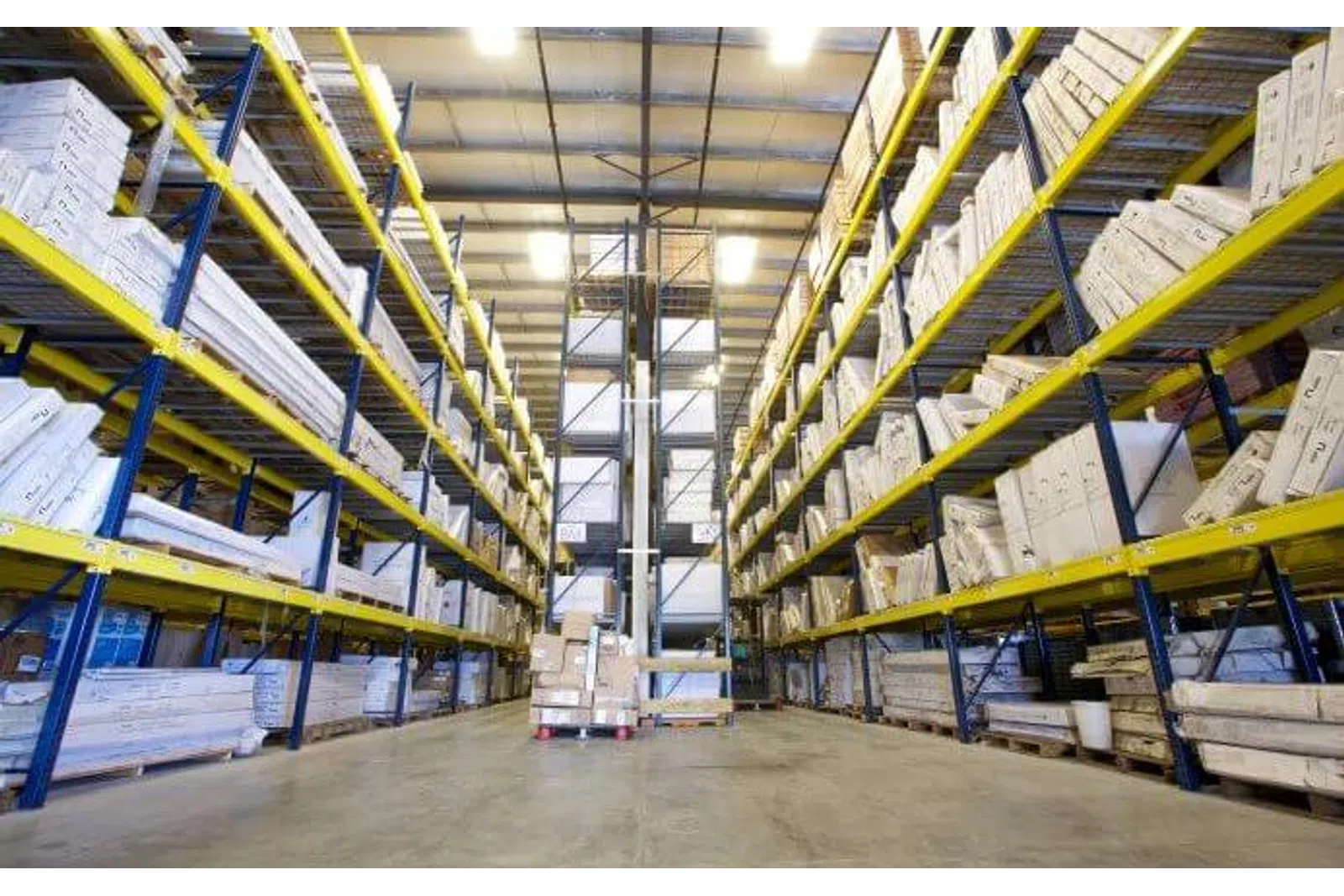 Large warehouse aisle with tall blue and yellow racks filled with stacked cardboard boxes and packages.