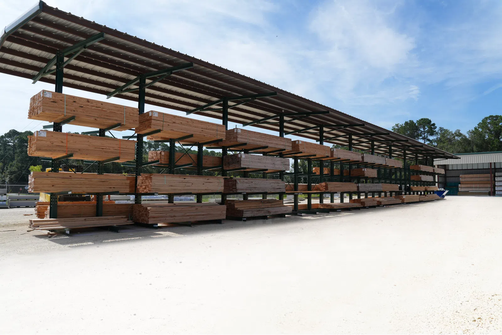 Outdoor lumber storage racks filled with wooden planks under a metal roof on a sunny day.