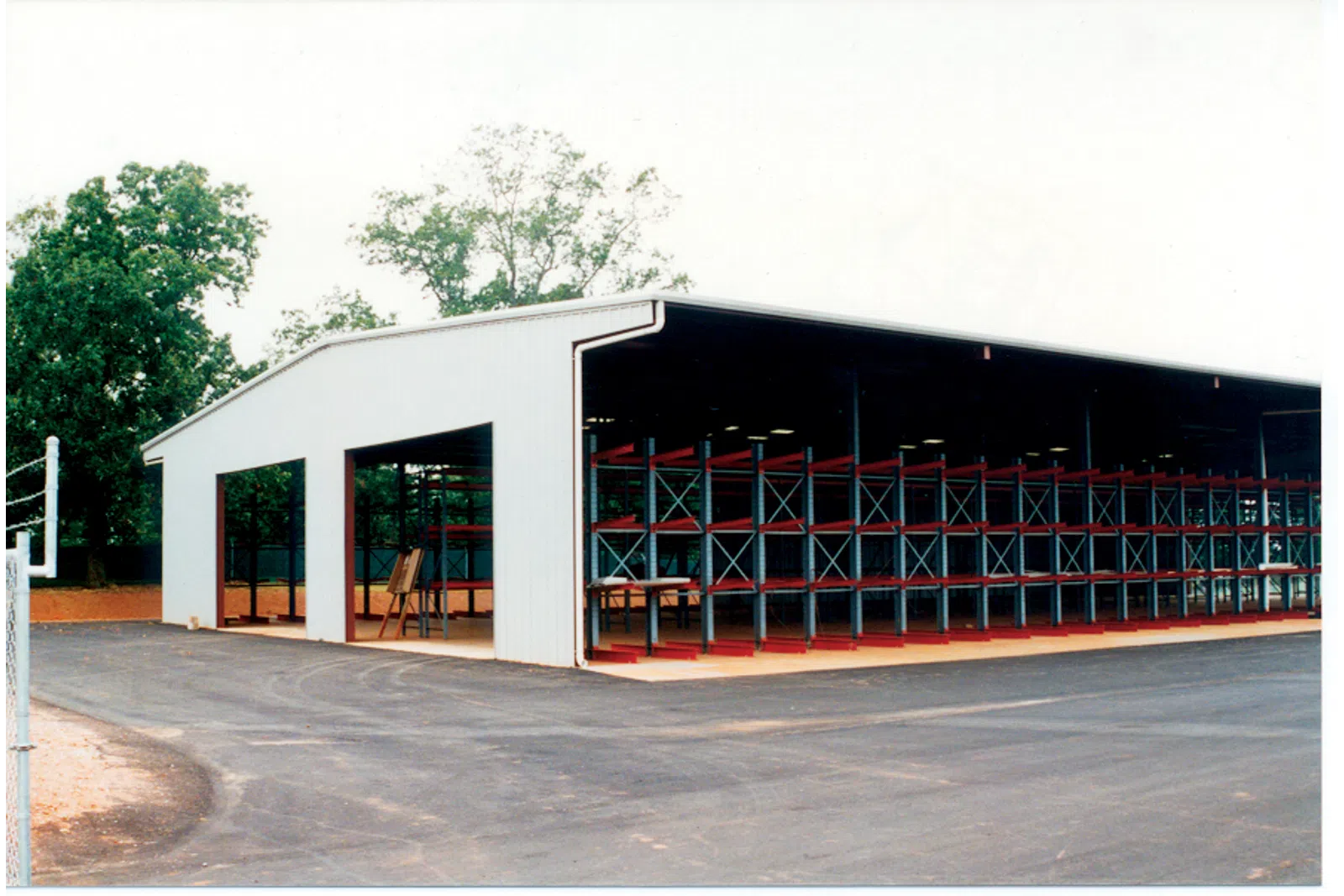 Large open warehouse with metal shelving units and paved driveway surrounded by trees on a clear day