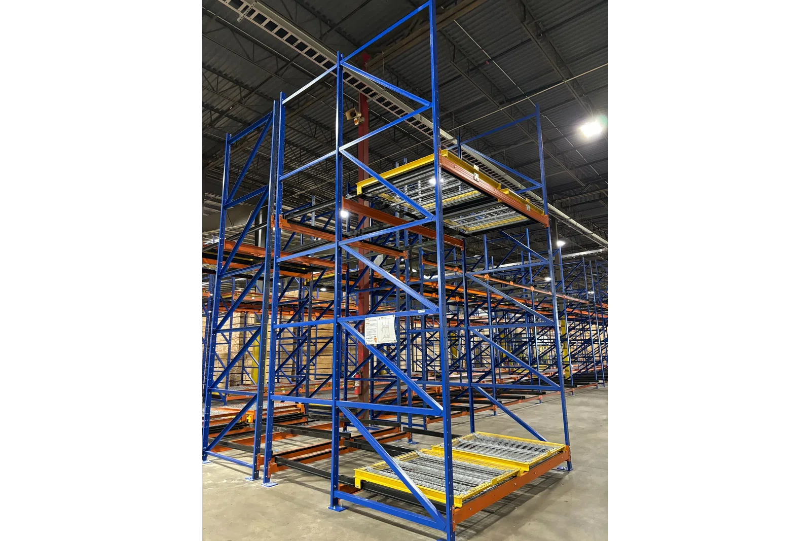 Empty blue and orange industrial warehouse shelving units under bright overhead lights on concrete floor.