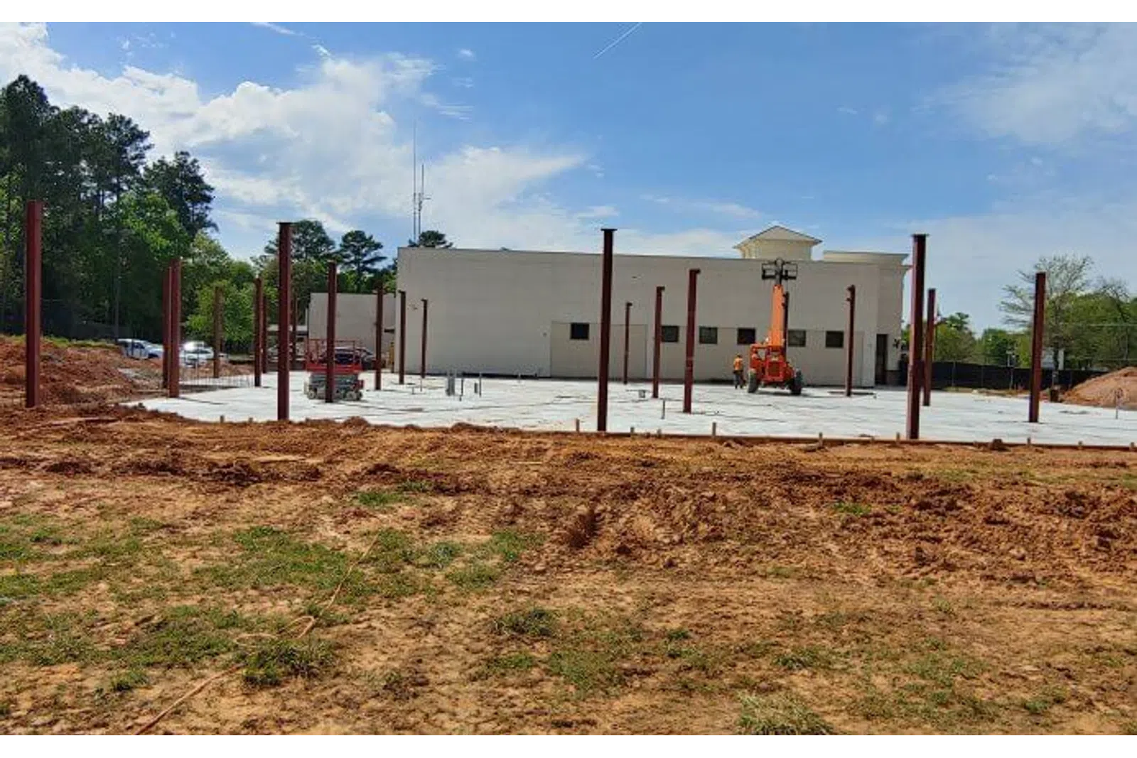 Construction site with steel beams installed on concrete foundation near a building under a blue sky.