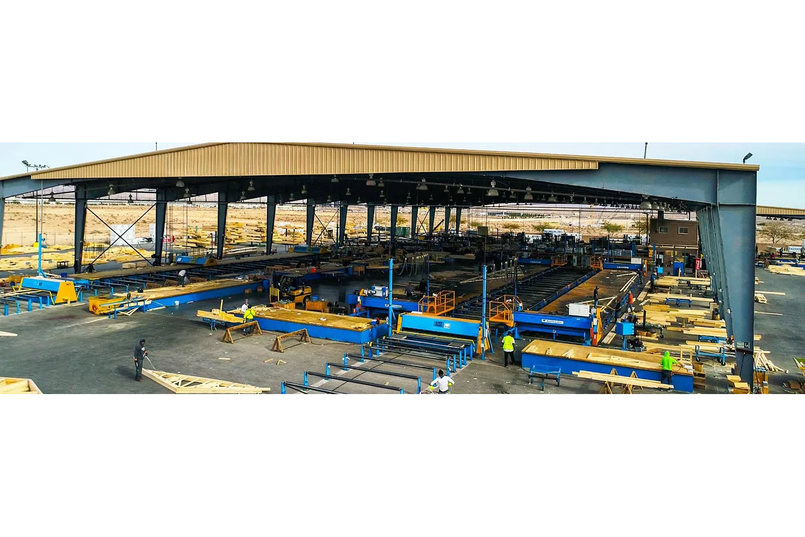 Wide view of an industrial metal fabrication workshop with workers and machinery under a large steel shed.