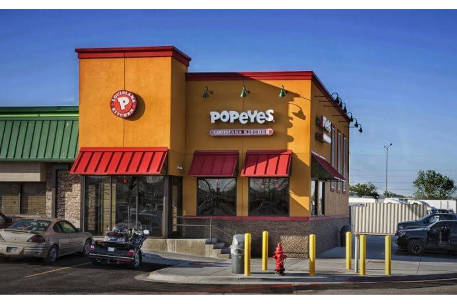 Exterior view of a Popeyes Louisiana Kitchen fast-food restaurant with bright orange and red facade under clear blue sky.