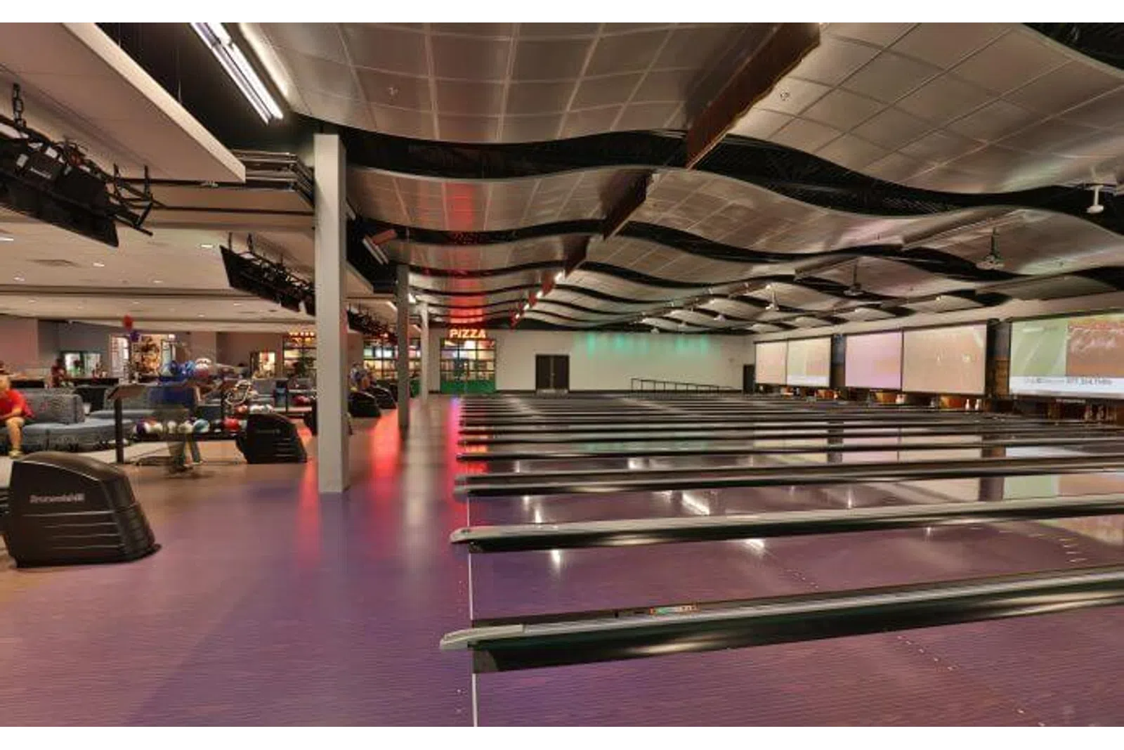Interior view of a spacious bowling alley with multiple lanes, seating areas, and a pizza counter in the back.
