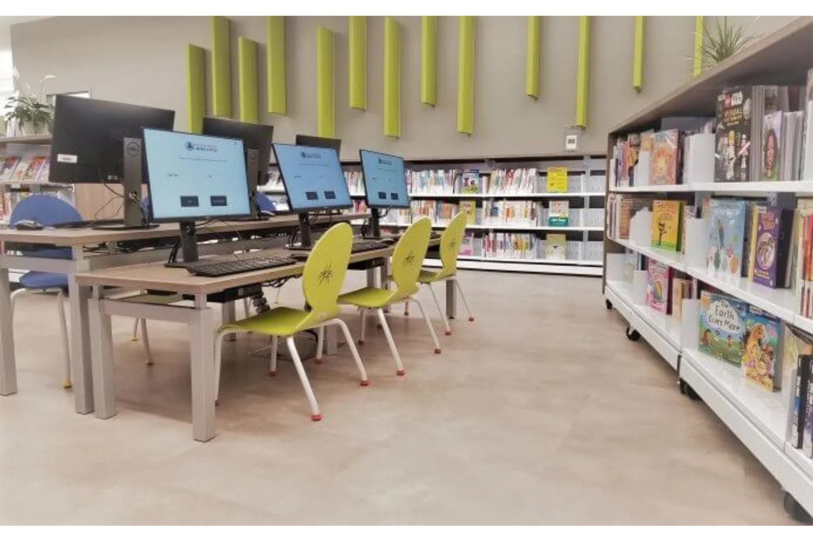 Library computer station with multiple monitors and green chairs beside bookshelves filled with books.