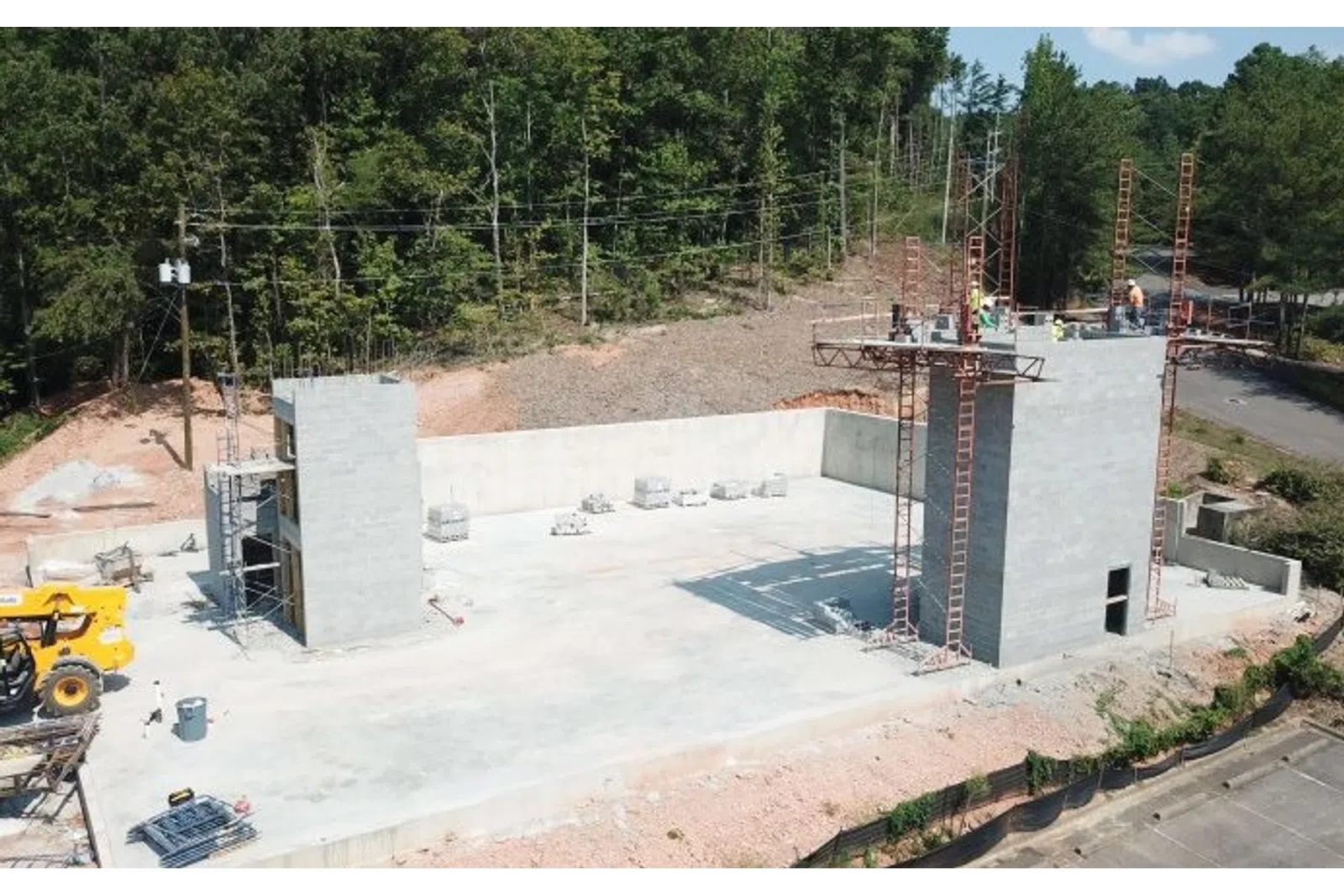 Construction site with concrete foundation and walls in early stages surrounded by trees and construction equipment.
