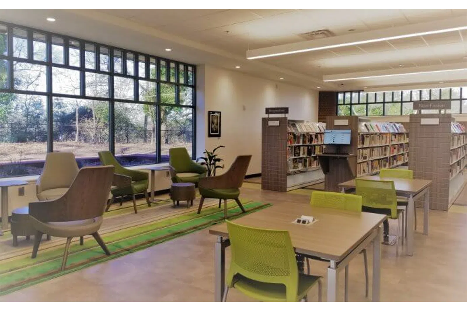 Modern library reading area with large windows, wooden tables, green chairs, and bookshelves under bright lighting.