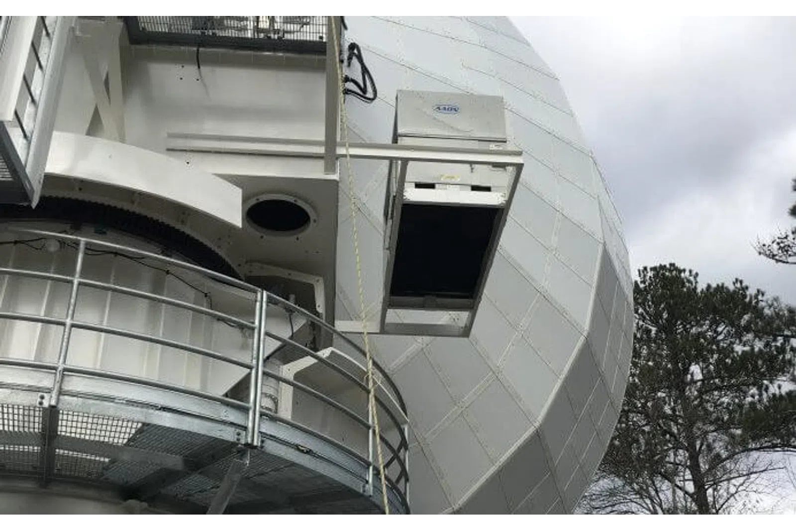Large white radar dome structure with metal platform railing and trees in the background under cloudy sky