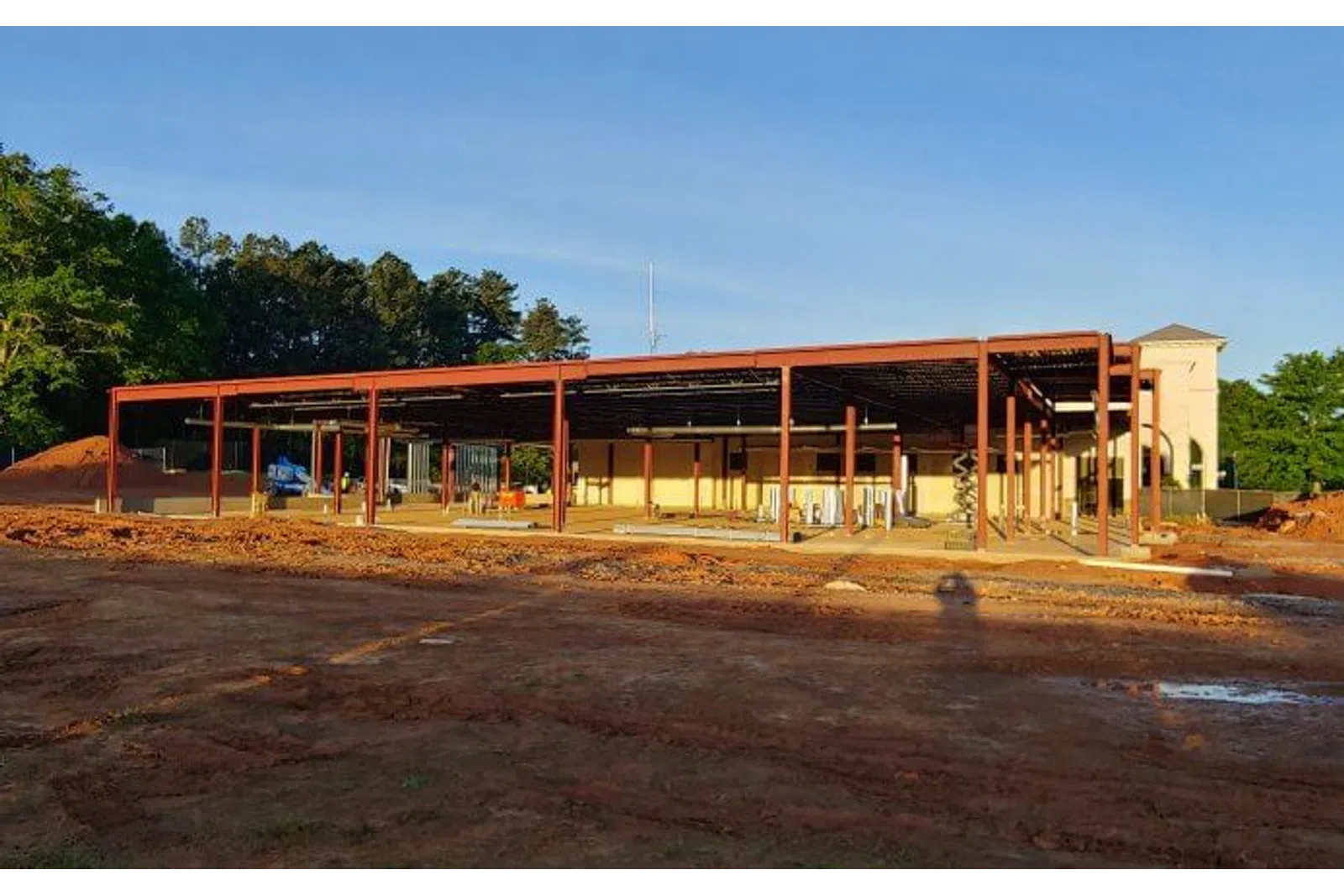 Steel framework of a building under construction on a cleared dirt site with trees and blue sky in the background