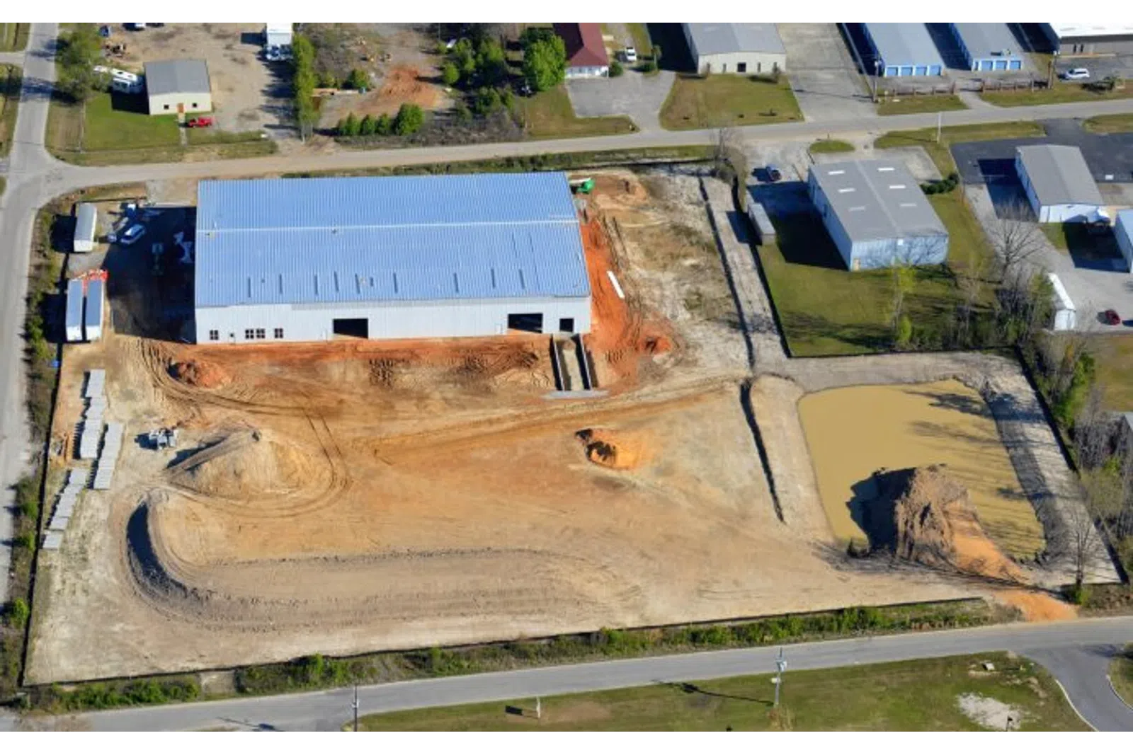 Aerial view of a construction site with a large warehouse, dirt roads, and a murky pond in an industrial area.