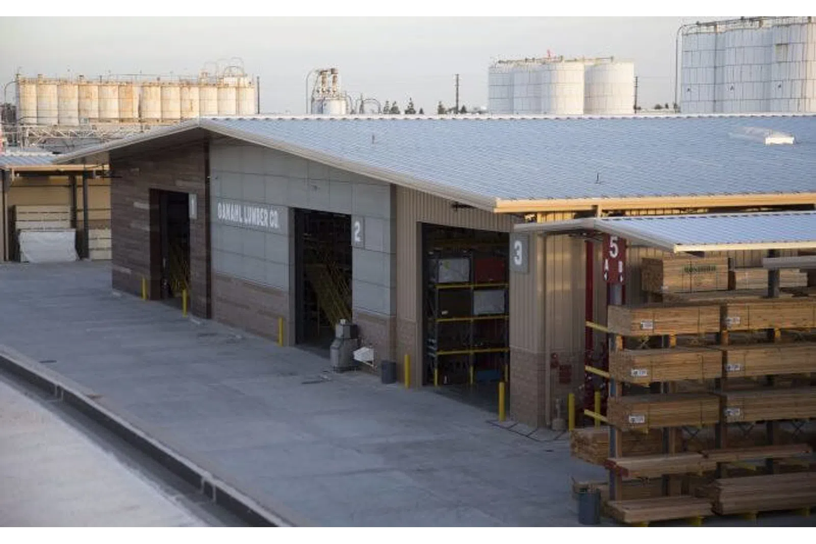 Outdoor lumber storage racks beside a warehouse building with industrial tanks in the background under a clear sky.
