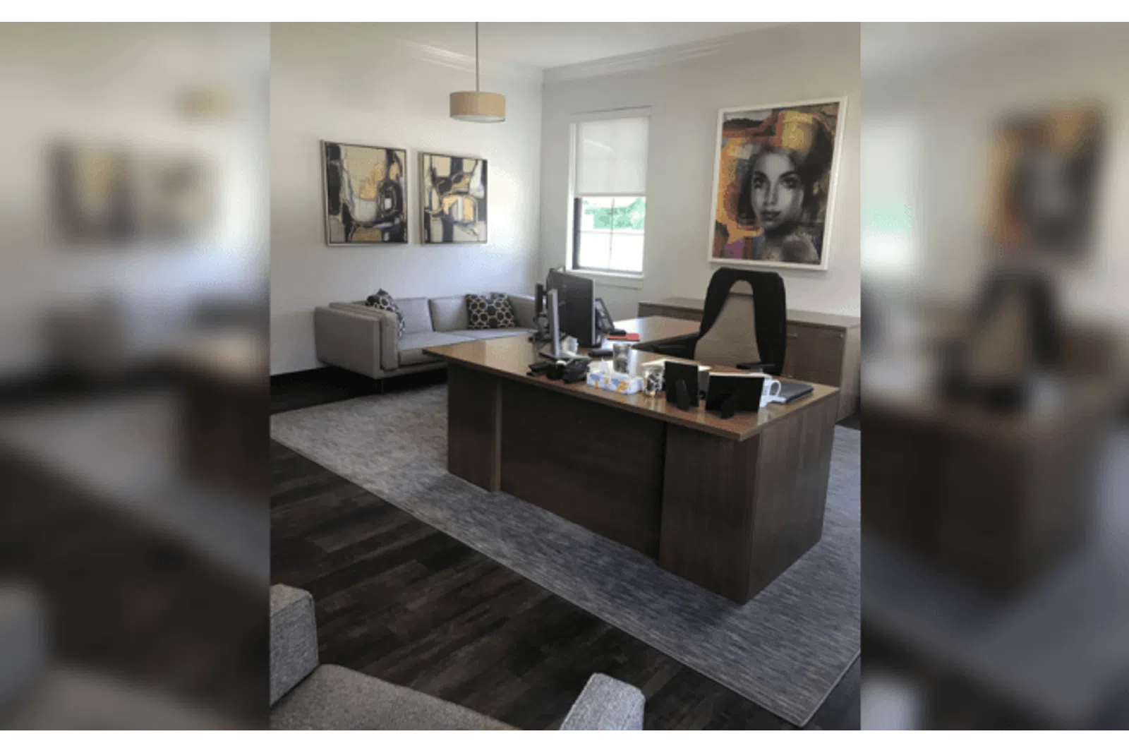 Modern office with wooden desk, black chair, couch, abstract art, and a large window with white walls.