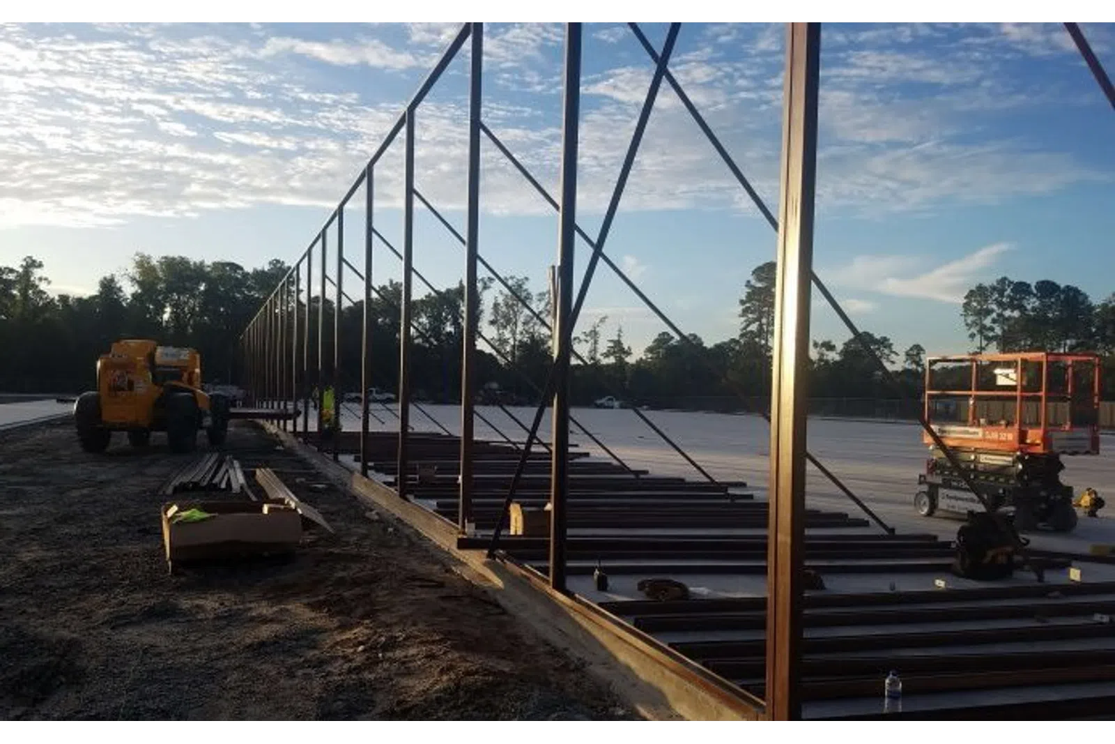 Steel framework under construction at a large outdoor site during early morning with construction equipment present