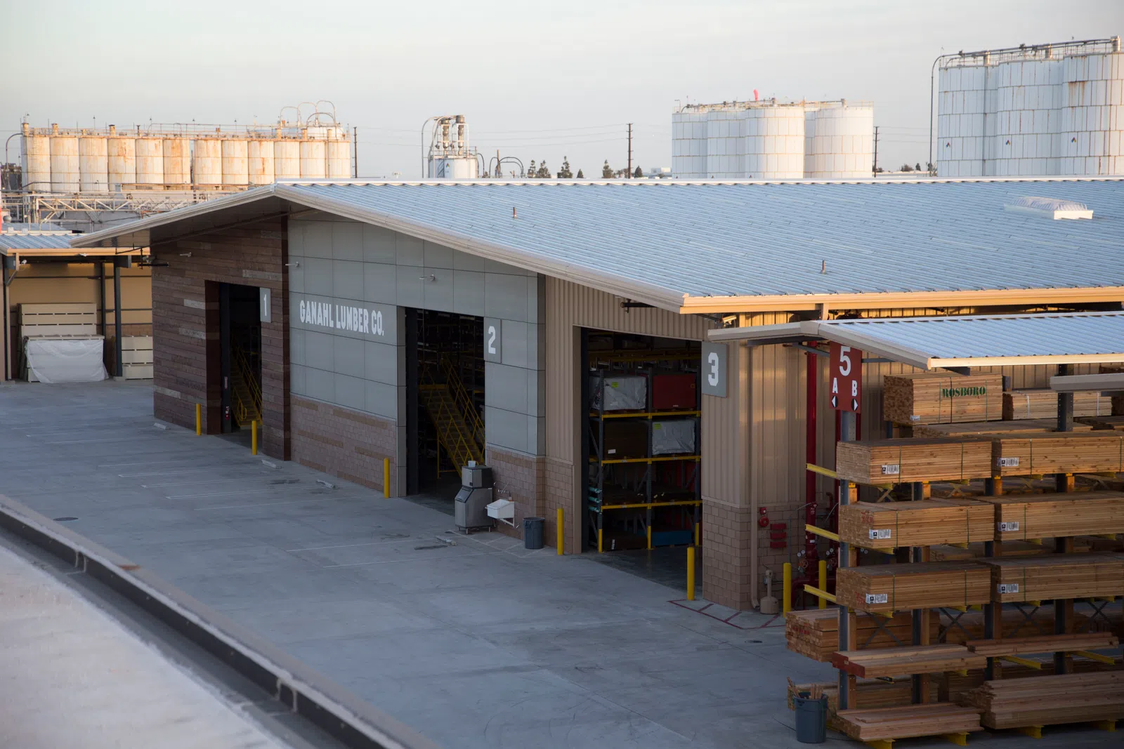 Warehouse exterior of Ganahl Lumber Co with stacked wooden planks and industrial tanks in background.