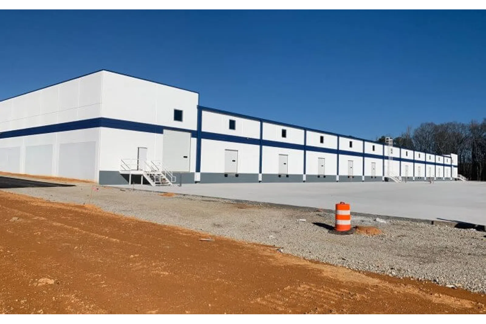 Large white industrial warehouse with a blue stripe under clear sunny sky, surrounded by dirt and gravel.