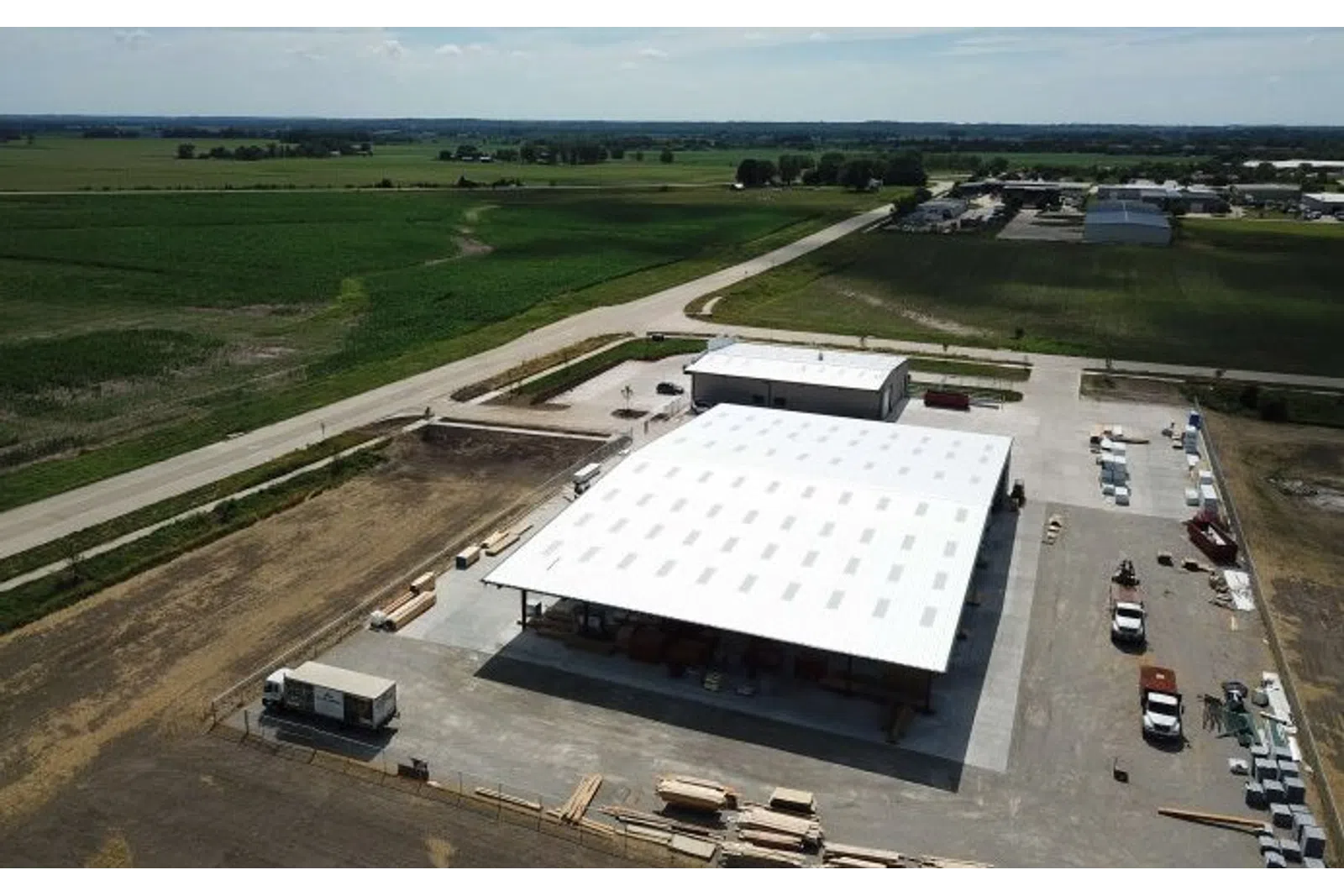 Aerial view of a large warehouse with white roof surrounded by farmland and vehicles on a sunny day