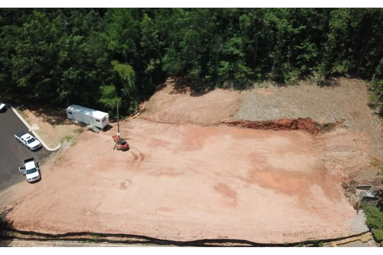 Aerial view of cleared construction site with dirt, parked vehicles, and surrounding trees in daylight.