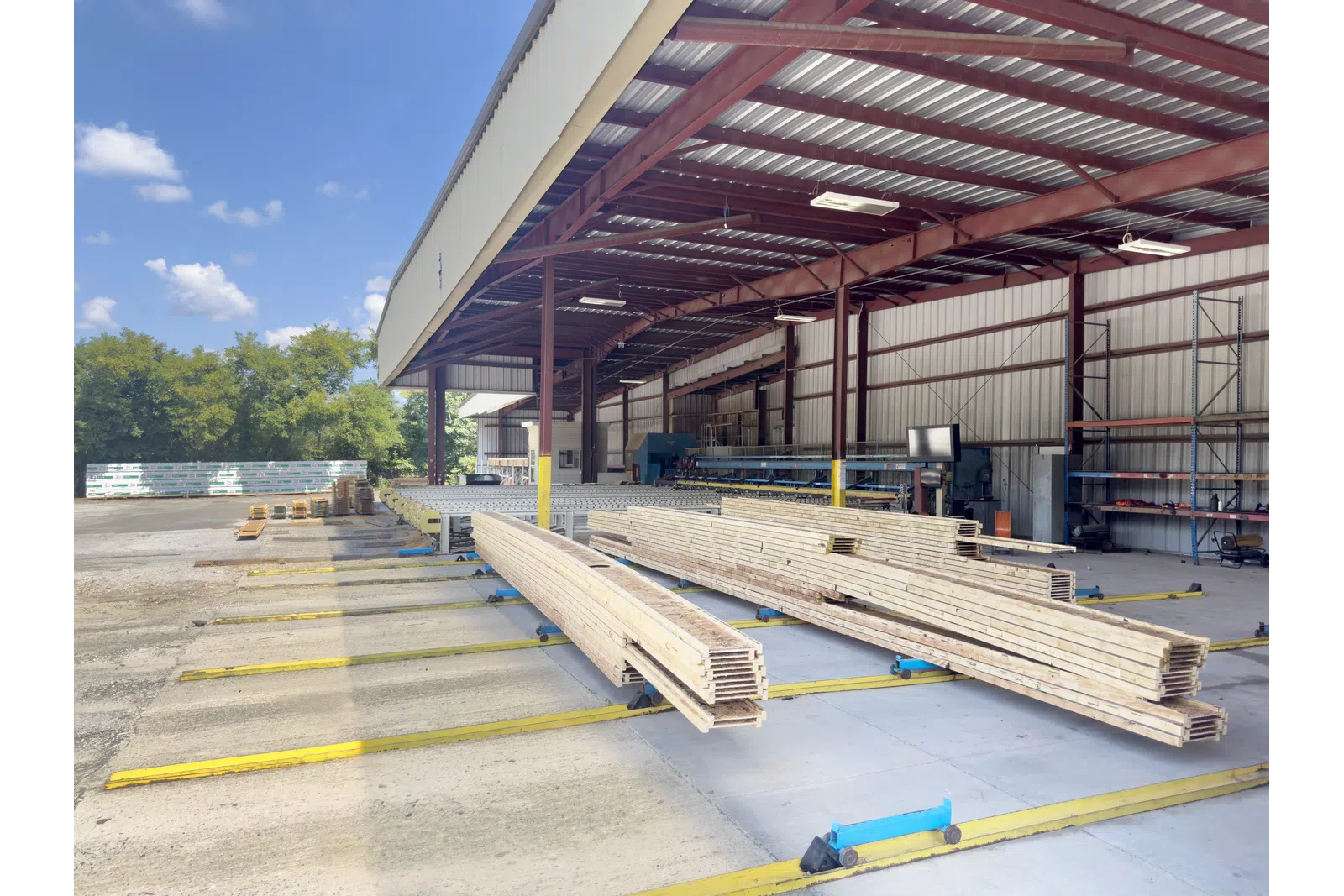 Stacked lumber under a metal roofed warehouse with clear sky and trees in the background