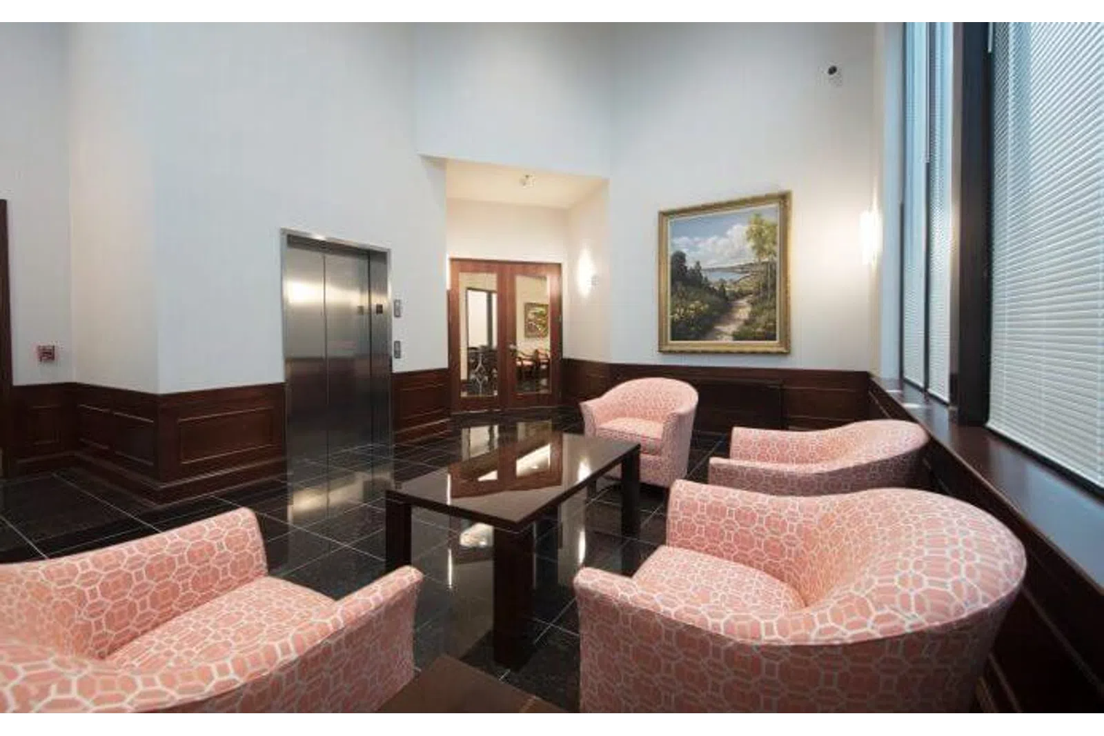 Lobby seating area with pink patterned chairs, glass coffee table, elevator, and framed landscape artwork.