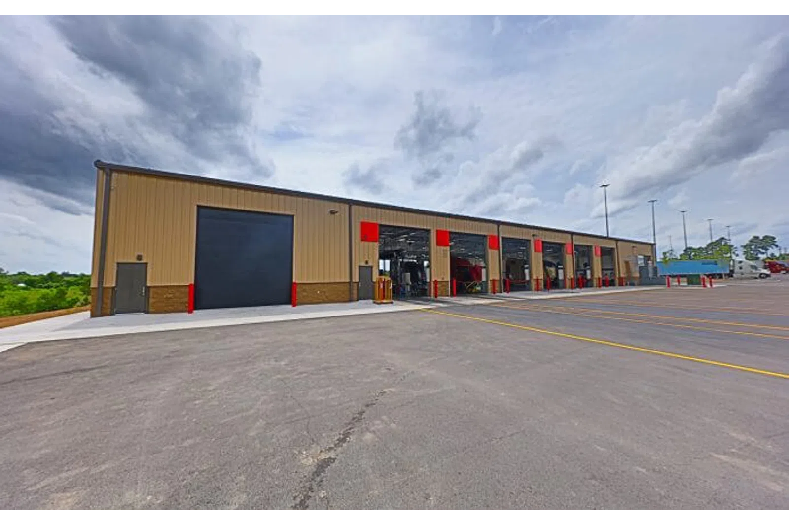 Wide view of a large industrial building with multiple open garage bays and a paved parking area under cloudy sky